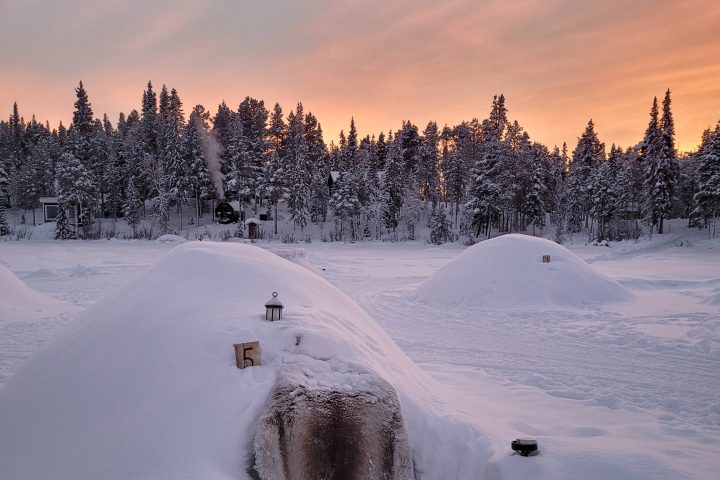 Snow-covered structures with forest and sunset sky in the background.
