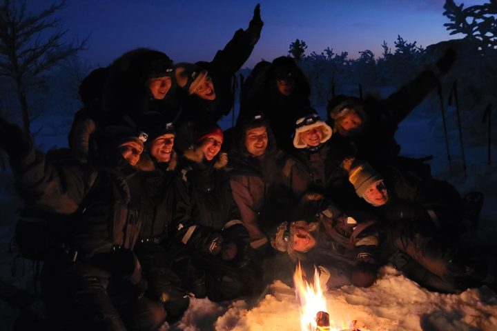 Group of people in winter gear around a campfire in a snowy landscape at dusk.