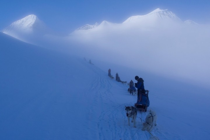Snowy landscape with sled dogs and people on a foggy mountain slope.