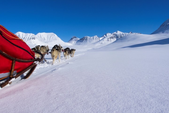 Sled pulled by dogs across snowy landscape with mountains under clear blue sky.