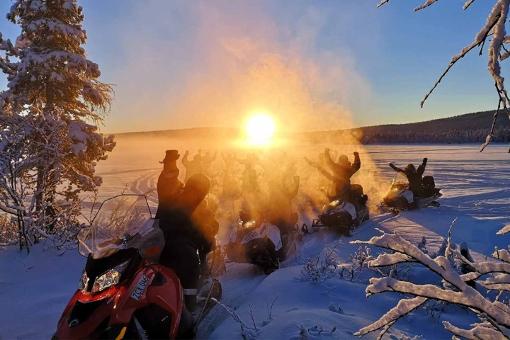 Snowmobilers celebrate at sunset on a snowy landscape with frosty trees.