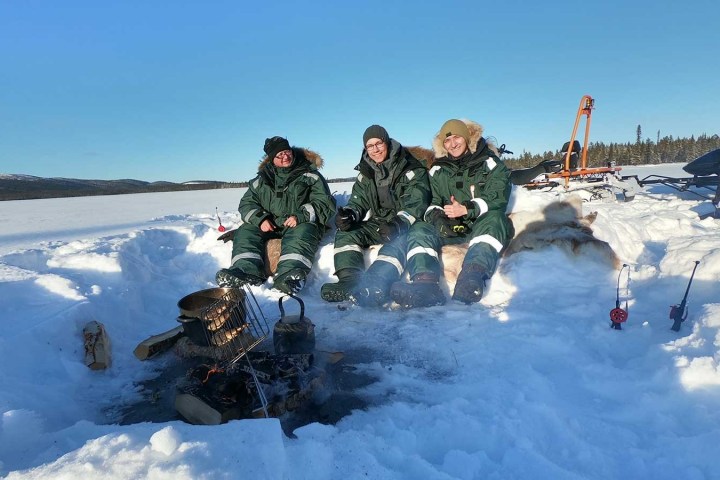 Three people in winter gear sit on snow by a campfire, with ice fishing rods and snowmobiles visible.