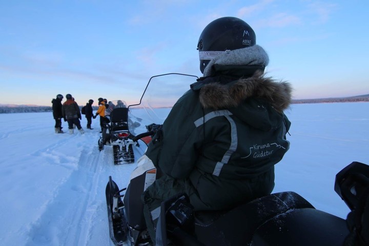 Person on snowmobile in winter gear with group in background on snowy landscape.