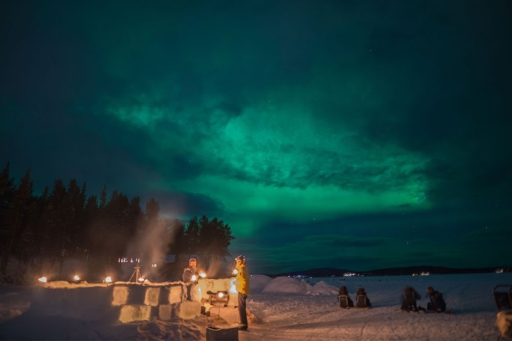 Aurora Borealis over snowy landscape with people near an ice bar and sleds.