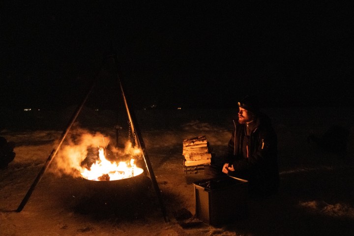 Person sitting by a campfire in the dark with snow and logs nearby.