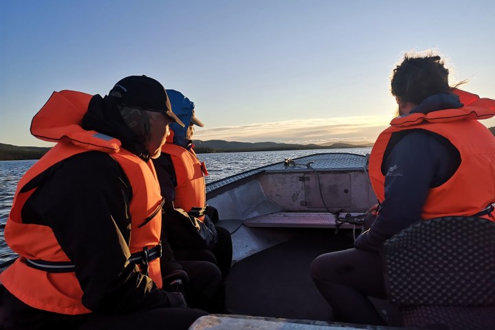 Three people in orange life vests sitting in a boat on a lake at sunset.