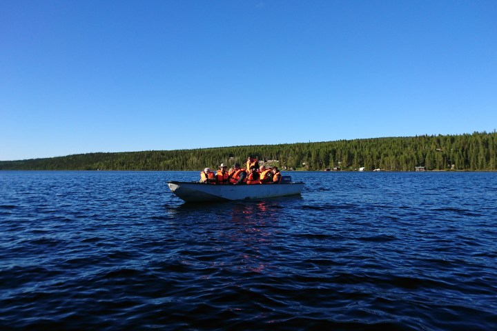 Boat with people wearing life jackets on a lake with forested shoreline under clear blue sky.