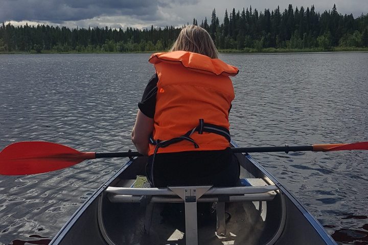 Person in life vest paddling a canoe on a calm lake, cloudy sky above.