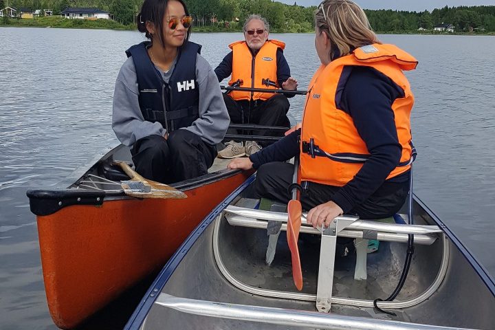 Three people wearing life jackets in canoes on a lake, with cloudy sky.