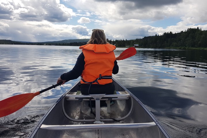 Person in an orange life vest paddling a canoe on a calm lake under cloudy sky.