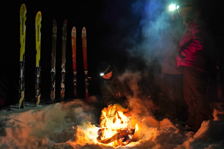 People around a campfire in the snow at night, with skis and headlamps.