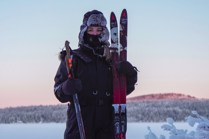 Person in winter clothing holding skis and poles against a snowy landscape at sunset.