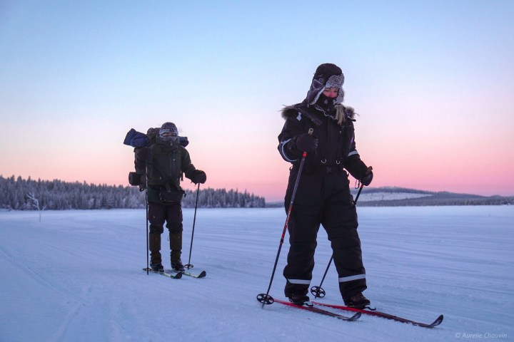 Two people skiing on snow with backpacks, against a pink sunset sky.