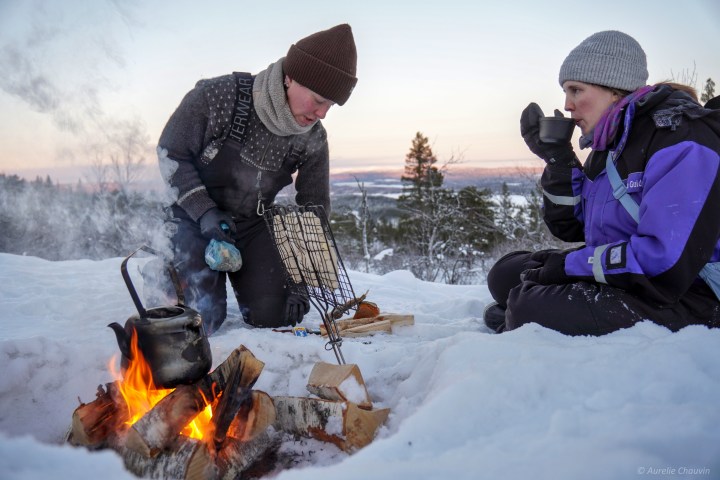 Two people enjoy a campfire in snowy landscape, one cooking, the other drinking from a mug.