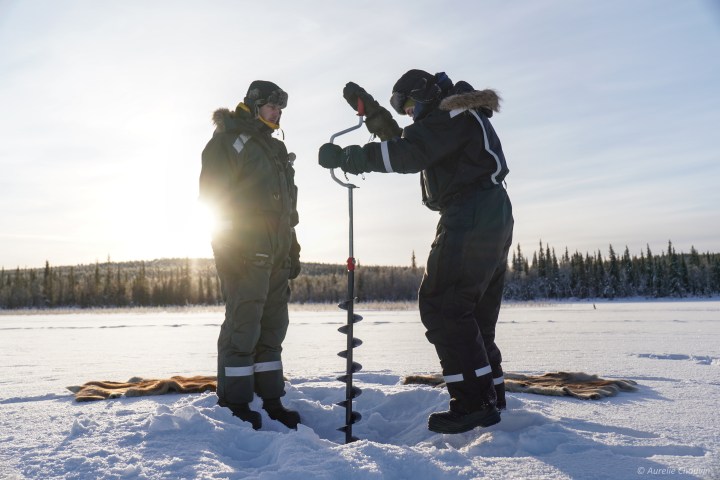 Two people drilling ice on a snowy landscape with trees in the background under a clear sky.