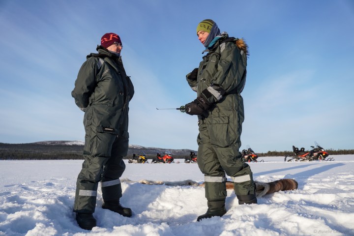 Two people in winter gear stand on snow, with snowmobiles and a scenic background.
