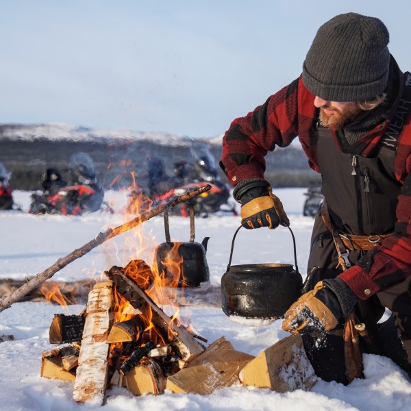 Person in winter gear kneeling by a campfire on snow, holding a kettle over the flames, snowmobiles in background.
