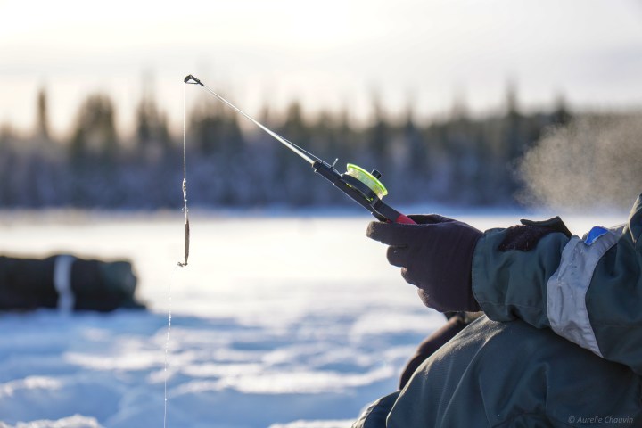 Person wearing gloves ice fishing on a frozen lake with a short fishing rod.