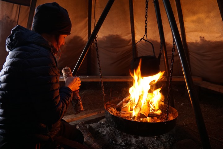 Person in warm clothes sitting by indoor fire, holding a log.