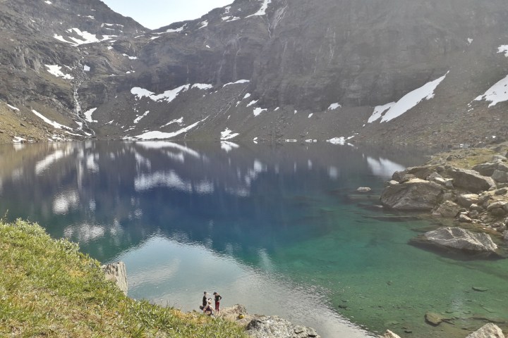 A clear mountain lake with rocky shores and patches of snow, reflecting cloudy sky and surrounded by rugged peaks.