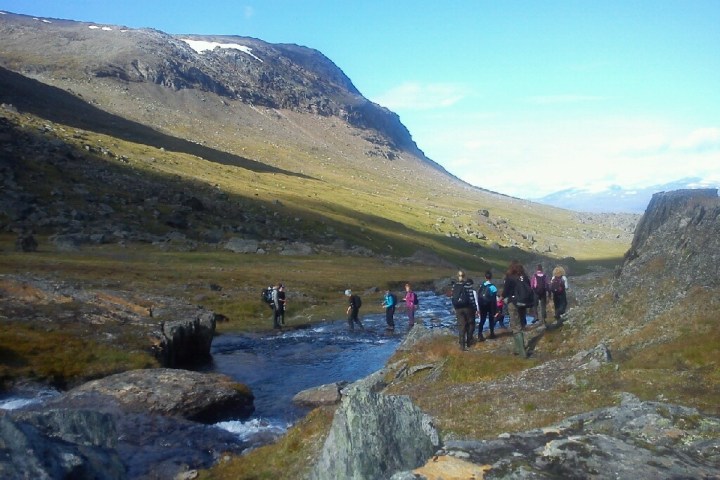 Group hiking through mountain terrain with a stream under a clear blue sky.