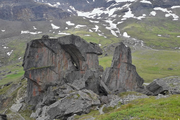 Large rocky formations in a mountainous landscape with patches of snow and green grass.