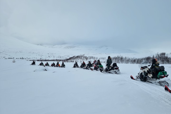 Line of snowmobiles with riders on a snowy landscape under cloudy skies.