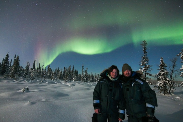 Two people in winter clothes under green aurora borealis in snowy forest.
