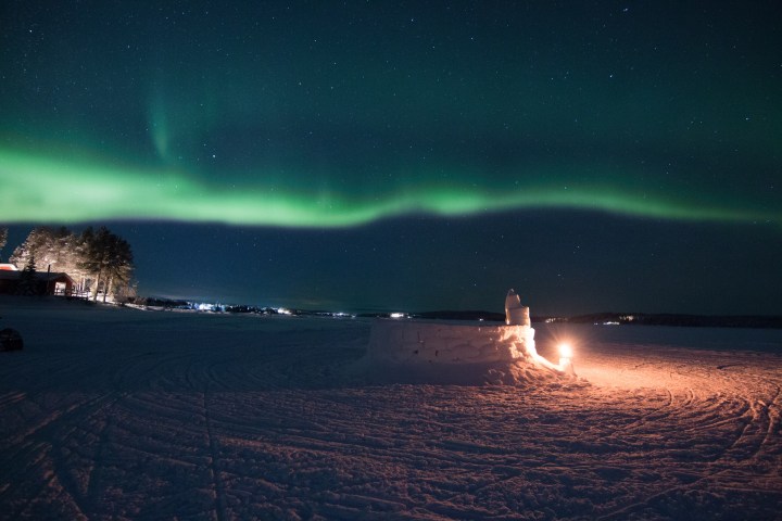 Northern lights over a snowy landscape with a lit igloo and distant trees at night.