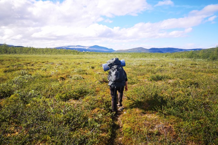 Backpacker with gear walking through a lush green meadow towards distant mountains under a blue sky.
