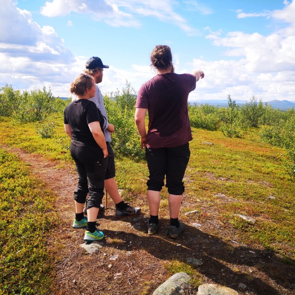 Three people standing on a grassy hill, one pointing to the distance under a partly cloudy sky.