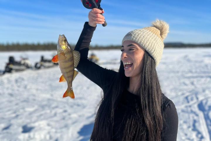 Person holding a fish they caught while ice fishing, wearing a beanie and smiling on a snowy landscape.