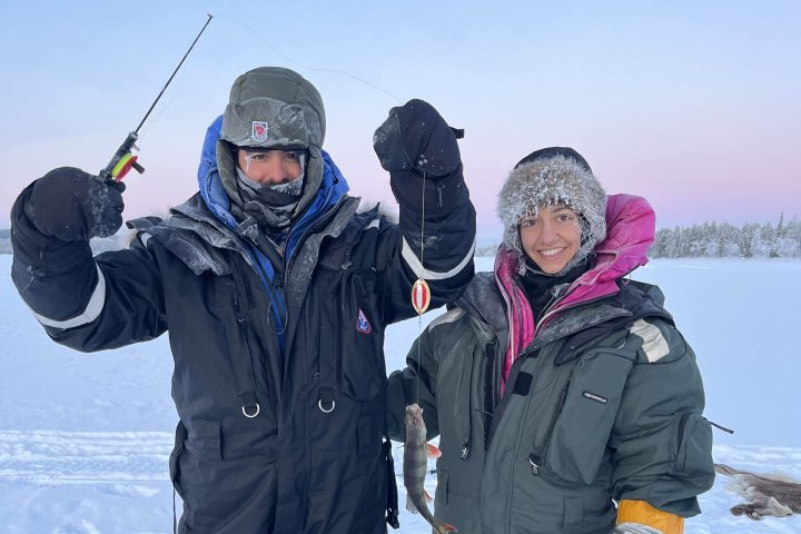 Two people in winter gear ice fishing, one holding a fish in snowy landscape.