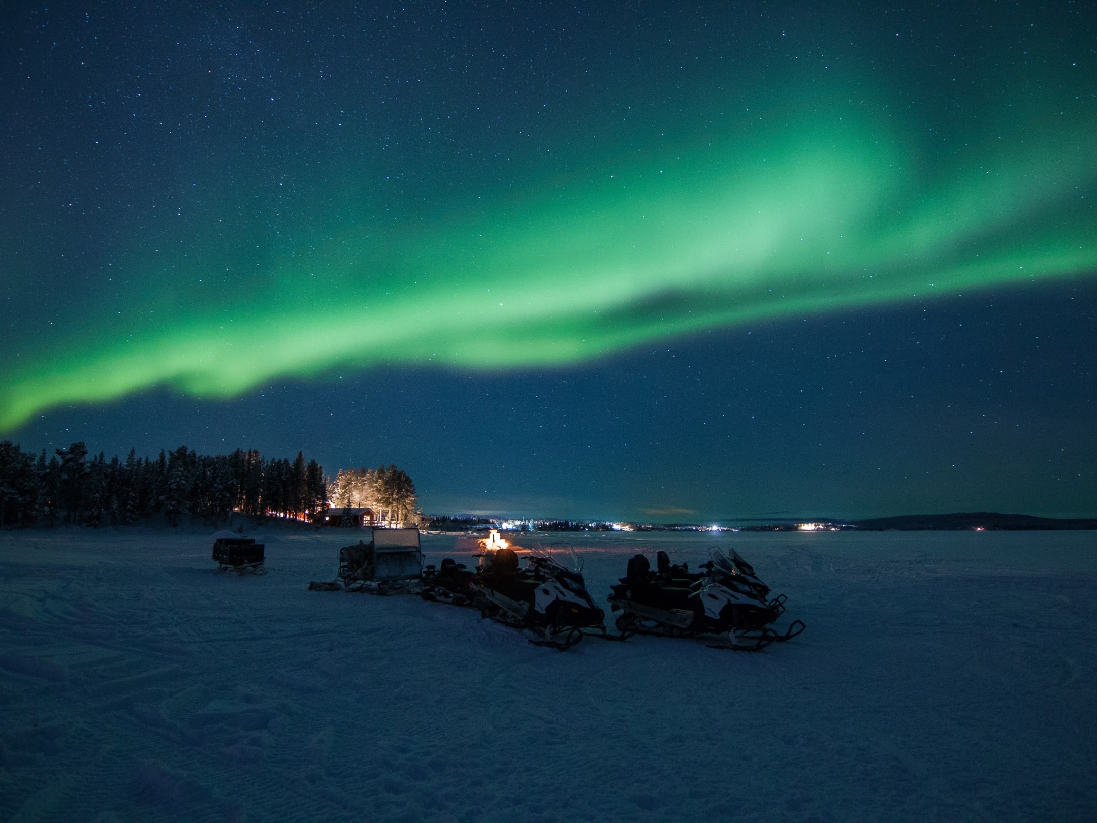 Northern lights over snowy landscape with snowmobiles in foreground.