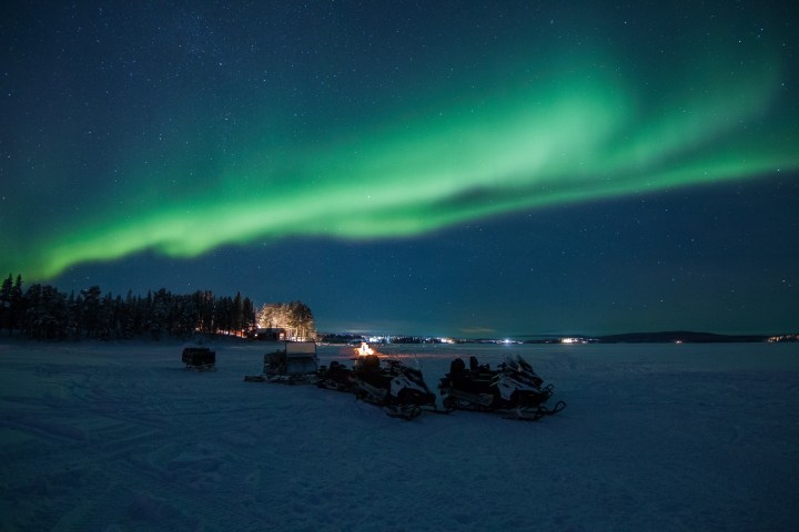 Northern lights over snowy landscape with snowmobiles in foreground.
