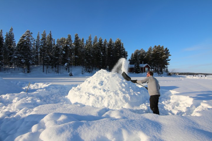 Person shoveling snow in a snowy landscape with trees and a house in the background.