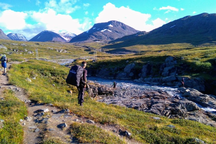 Hikers on a rocky path near a river with mountains in the background under a blue sky.