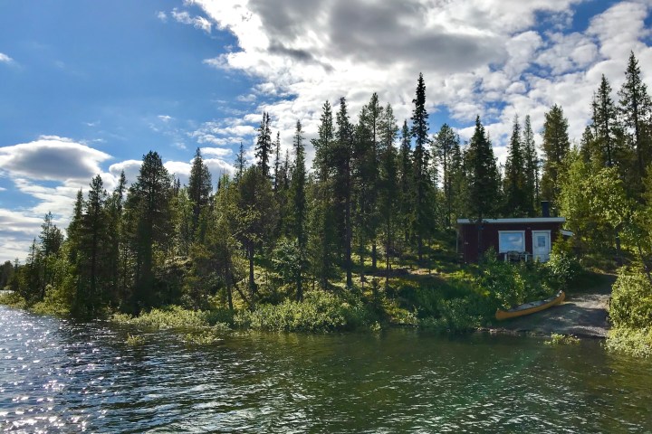 Lakeside cabin surrounded by trees under a partly cloudy sky, with a canoe on the shore.