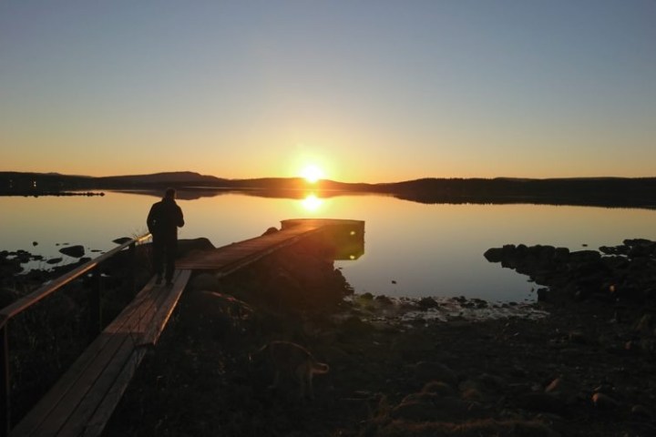 Person on boardwalk over calm lake at sunset, clear sky with distant hills.