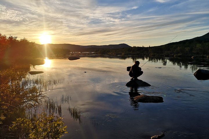 Silhouette of a person fishing on a rock by a lake at sunset.