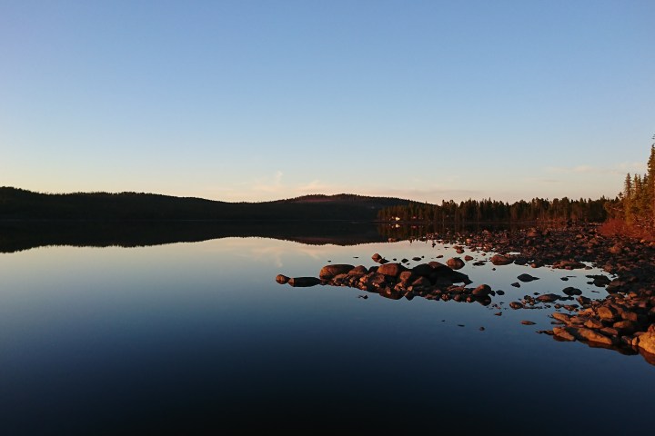 Calm lake reflecting sky and tree-lined shore at sunset with rocks in foreground.