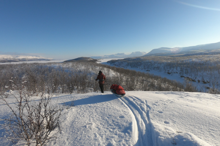 Person pulling sled through snowy landscape with mountain views and clear blue sky.
