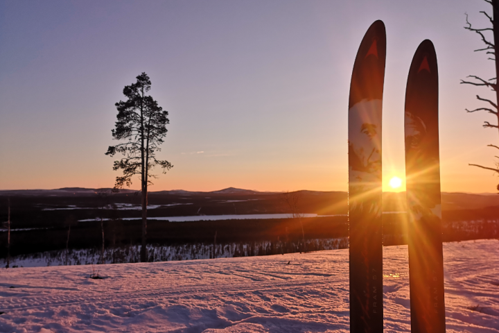 Sunset over snowy landscape with two skis, tree silhouette, and distant hills.