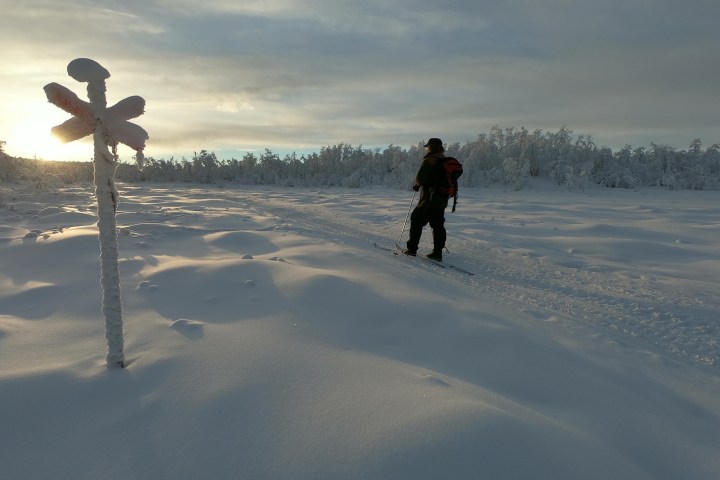 Person skiing on snow-covered landscape with a snowy signpost in foreground during sunset.