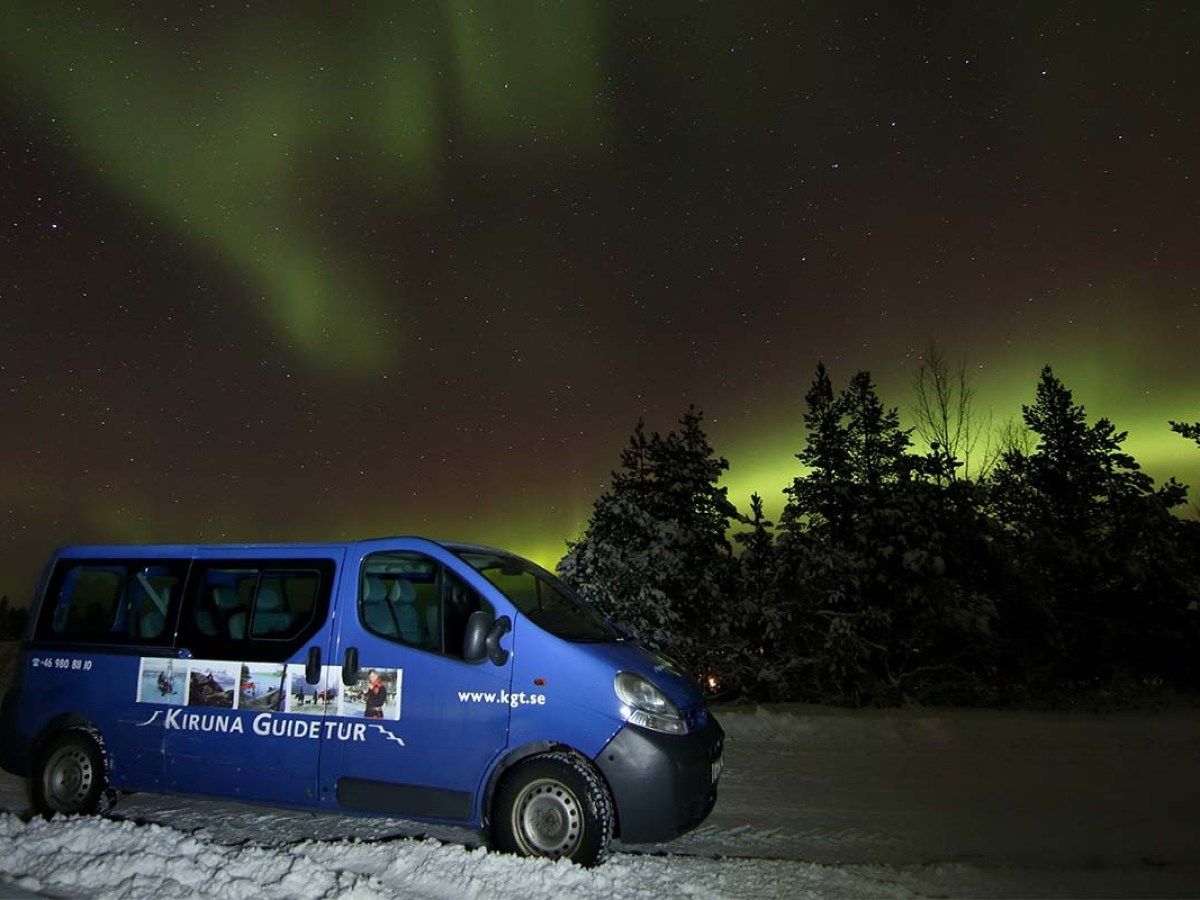 Blue tour van parked on snowy ground with northern lights in the sky.
