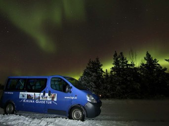 Blue tour van parked on snowy ground with northern lights in the sky.