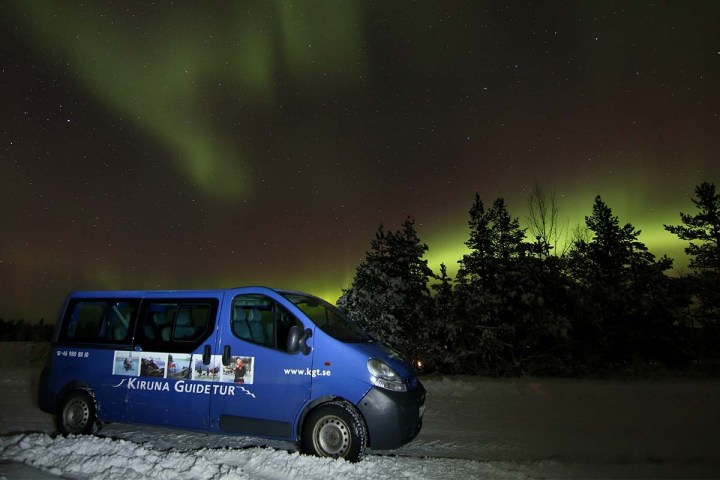 Blue tour van parked on snowy ground with northern lights in the sky.