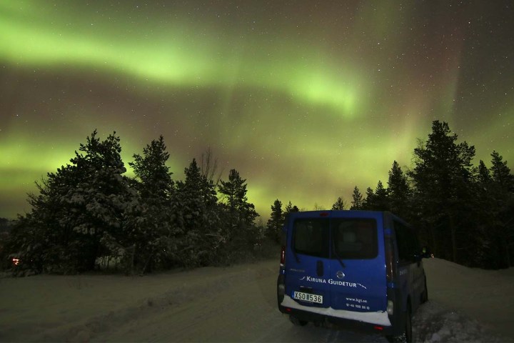 Blue van on snowy road under green aurora borealis with pine trees.