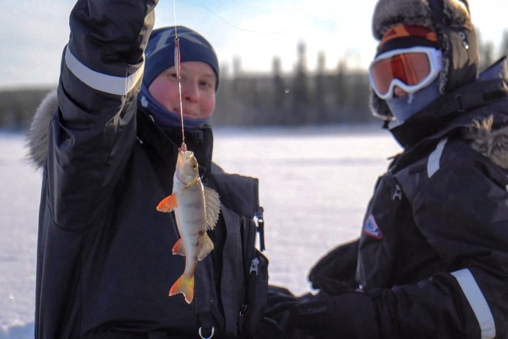 Two people in winter gear, one holding a fish on a line during ice fishing.