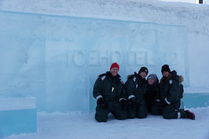 Four people in winter gear kneeling in front of an ice hotel entrance.